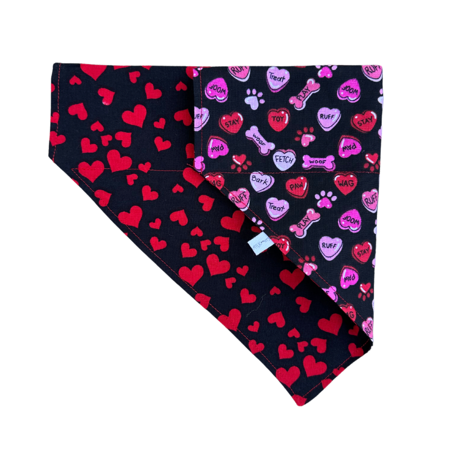 A reversible dog bandana with one side featuring a black fabric with red hearts and the other side featuring a black fabric with dog-themed conversation hearts.