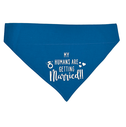 A blue dog bandana with white text 'MY HUMANS ARE GETTING MARRIED' printed on it, along with a graphic of a ring and hearts.