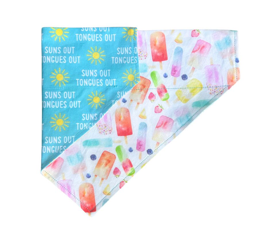 A folded fabric bandana or scarf featuring a split design: one side is light blue with yellow suns and the text 'SUNS OUT TONGUES OUT,' and the other side is white with colorful watercolor popsicle prints.
