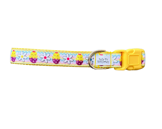 A colorful dog collar with a pattern of chicks, eggs, and daisies, predominantly yellow and purple, with a yellow plastic buckle.